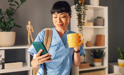 Nurse in blue scrubs drinking coffee and using her phone at home